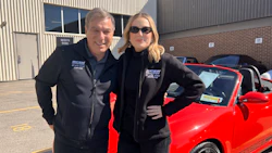 Two automotive professionals in black jackets posing with a red sports car