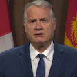 Government official speaking at press conference with Canadian provincial flags