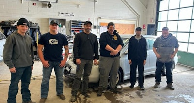 Auto repair team of six workers standing together in garage workshop beside white car