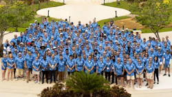 Large professional team in blue shirts gathered for group photo on outdoor landscaped pathway