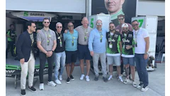 Racing team members gathered in paddock area wearing casual attire and sunglasses