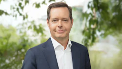 Professional man in navy suit and white shirt standing outdoors with blurred green foliage background