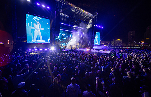 Nighttime music festival with large stage, video screens, and packed crowd under blue lighting