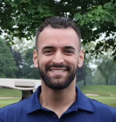 Smiling man in blue polo shirt standing outdoors with trees in background