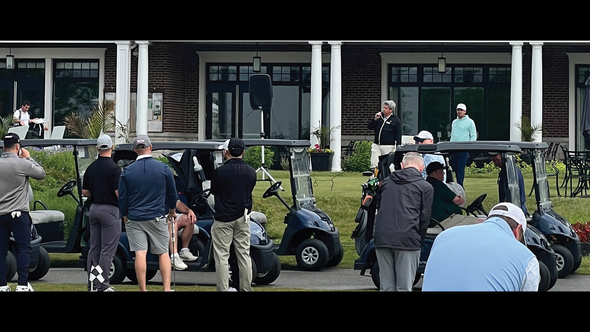 Golfers gathered with golf carts at outdoor event near brick building