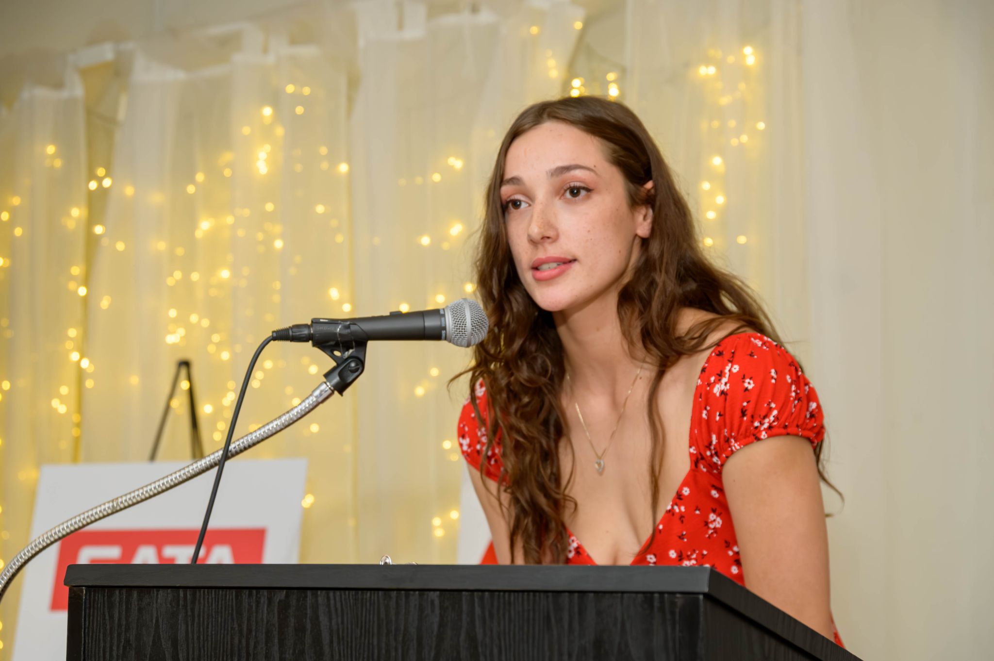 Woman in red floral dress speaking at a microphone with golden fairy lights backdrop