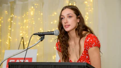 Woman in red floral dress speaking at a microphone with golden fairy lights backdrop