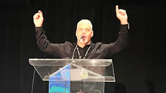 Speaker with raised arms addressing audience from a clear podium on dark stage