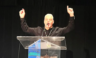 Speaker with raised arms addressing audience from a clear podium on dark stage