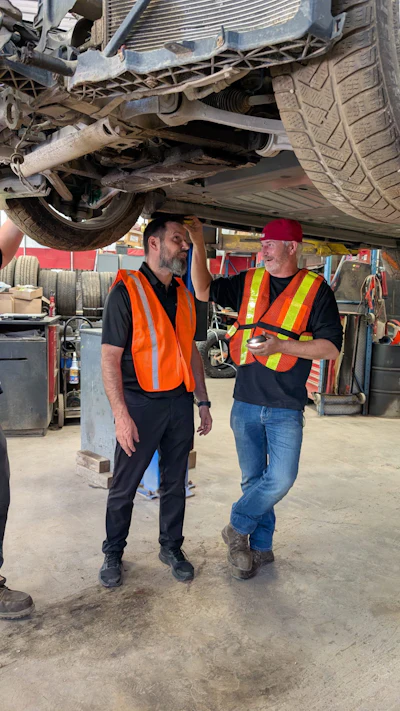 Two automotive technicians in safety vests inspecting vehicle undercarriage in workshop