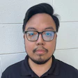 Professional headshot of a person wearing black-rimmed glasses and a dark polo shirt