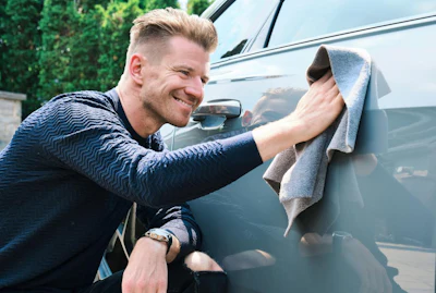 Smiling man wiping a silver car with a gray towel outdoors