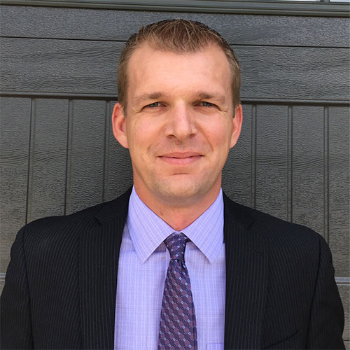 Professional man in black suit and purple shirt smiling against gray background
