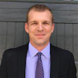 Professional man in black suit and purple shirt smiling against gray background