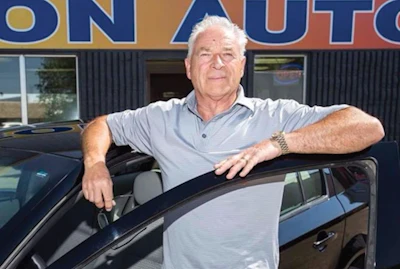 Mature man leaning on car door at auto dealership with blue storefront sign