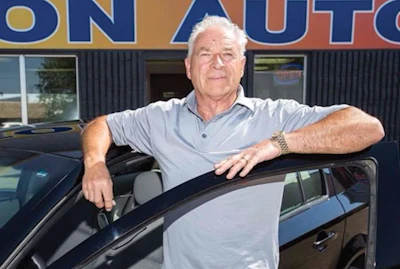 Mature man leaning on car door at auto dealership with blue storefront sign