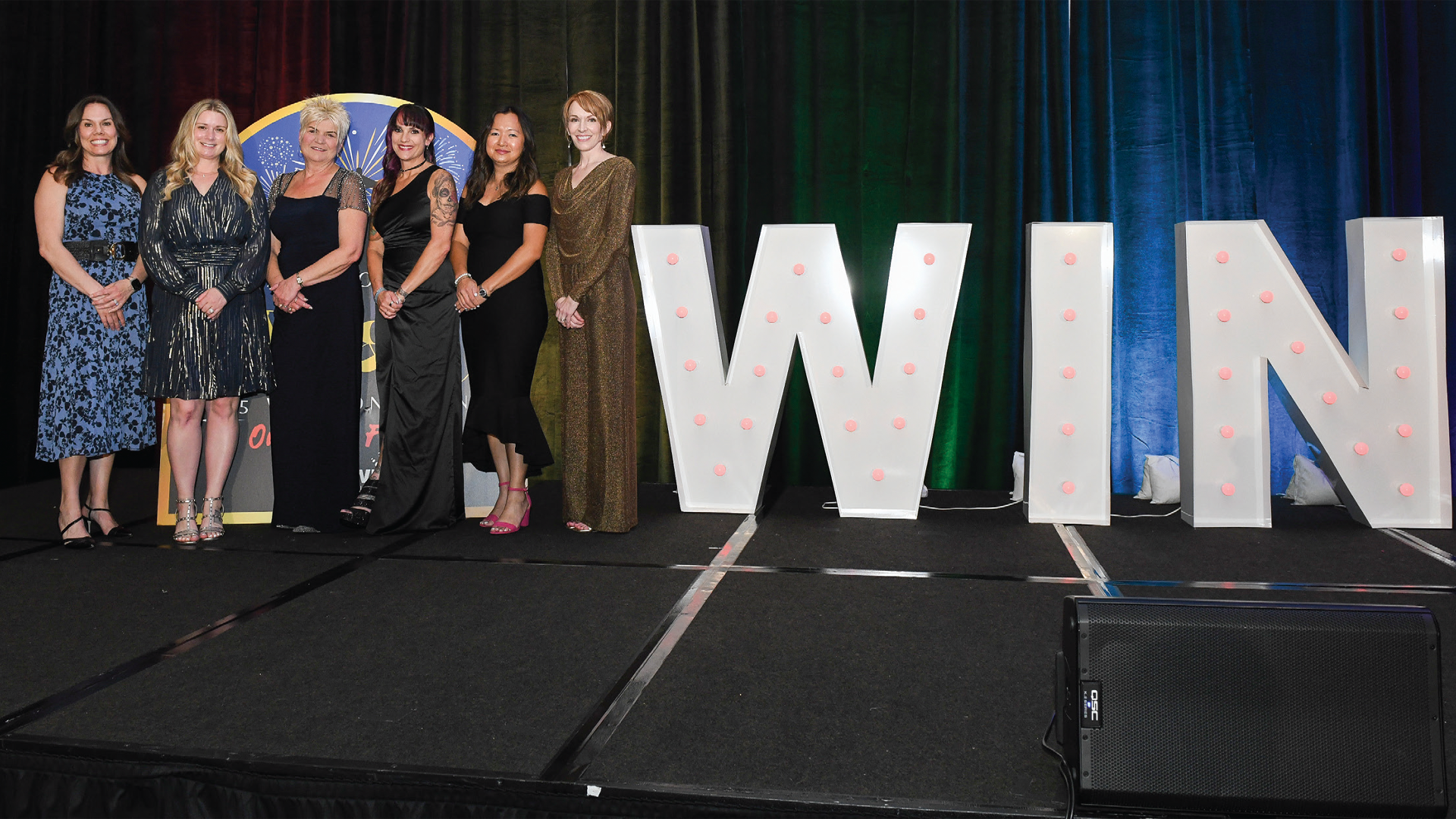 Six women standing on stage next to large illuminated WIN letters