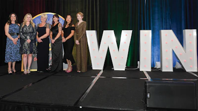 Six women standing on stage next to large illuminated WIN letters