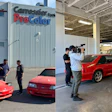 Red sports car inside auto body shop with camera crew and technicians