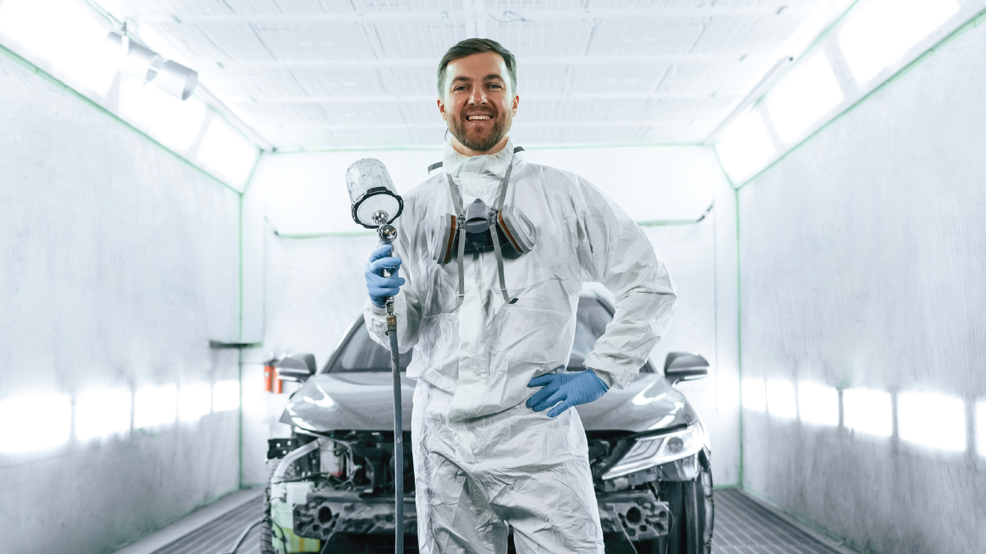 Auto painter in white protective suit smiling in bright spray booth with vehicle