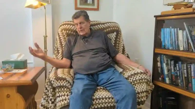 Middle-aged man in gray shirt and blue jeans sitting in a patterned armchair near bookshelf