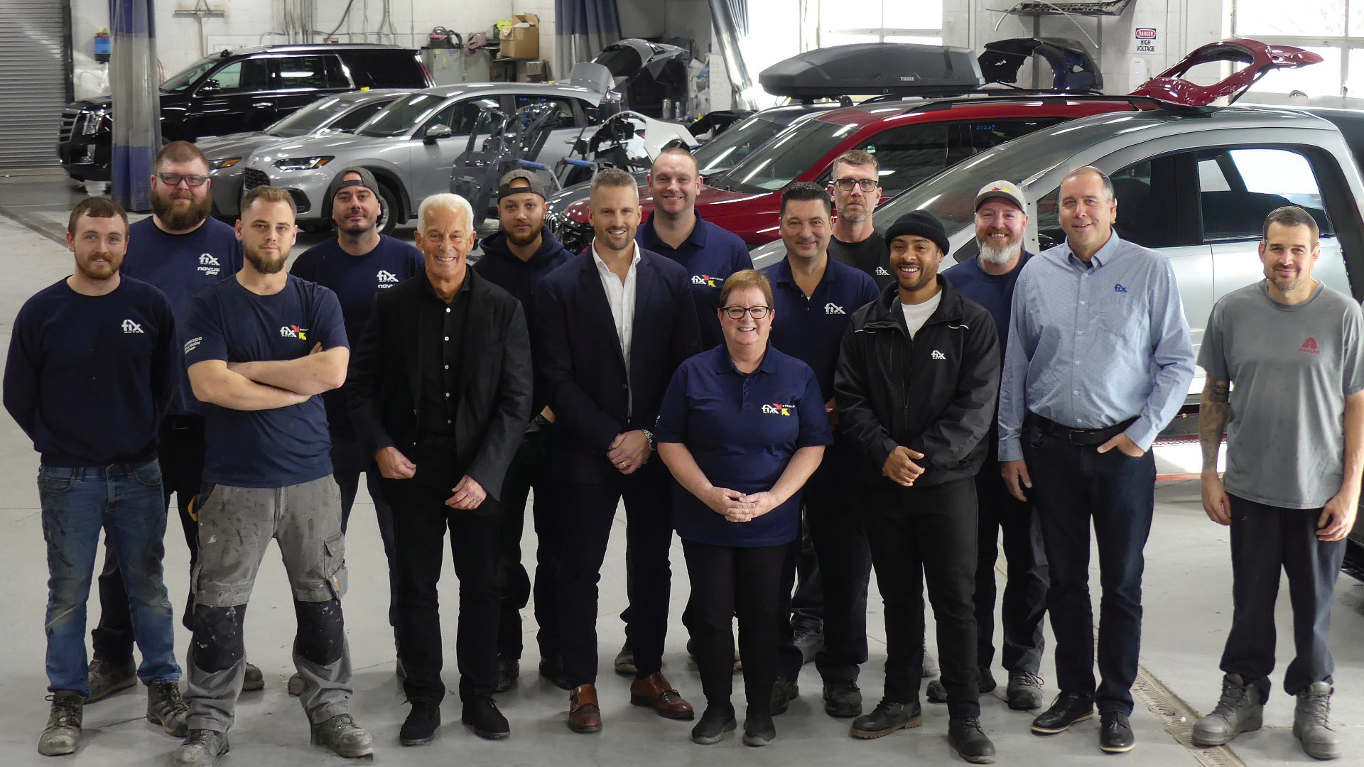 Automotive team of 15 professionals posing together in a car workshop with vehicles in background