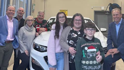 Group of eight people posing with a white vehicle in an indoor showroom