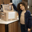 Young child smiling beside a cardboard battery recycling collection box in an indoor facility