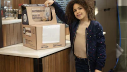 Young child smiling beside a cardboard battery recycling collection box in an indoor facility