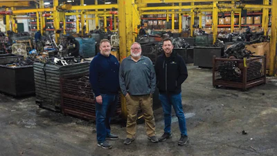 Three workers standing in industrial machine shop with metal equipment and yellow scaffolding