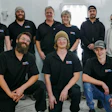 Team of nine professionals in black branded polos posing together indoors for a group photo