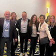 Eight professionals wearing conference badges pose together at a business event with modern lighting backdrop
