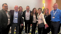 Eight professionals wearing conference badges pose together at a business event with modern lighting backdrop