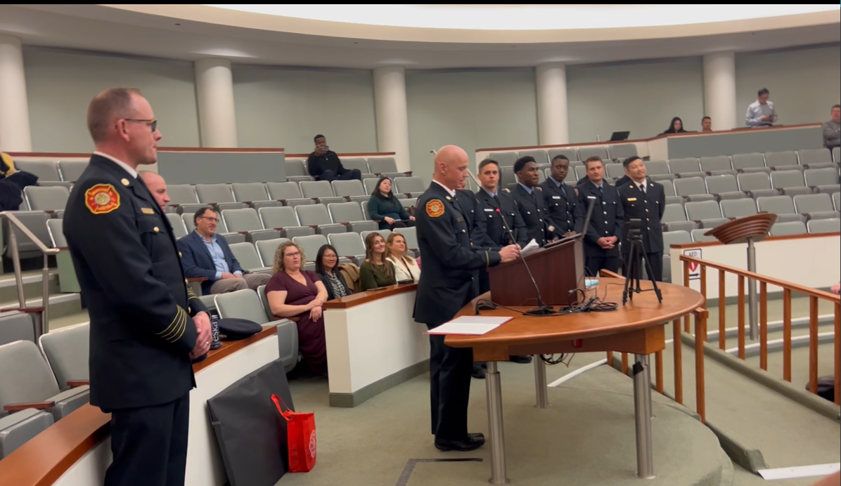 Law enforcement officials in dress uniforms stand at podium during formal ceremony in auditorium with seated attendees.