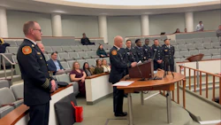 Law enforcement officials in dress uniforms stand at podium during formal ceremony in auditorium with seated attendees.