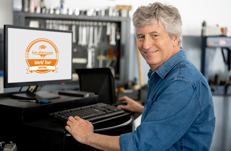 Man in denim shirt smiling at computer displaying Car-Part.com World Tour logo in workshop