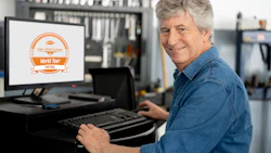 Man in denim shirt smiling at computer displaying Car-Part.com World Tour logo in workshop