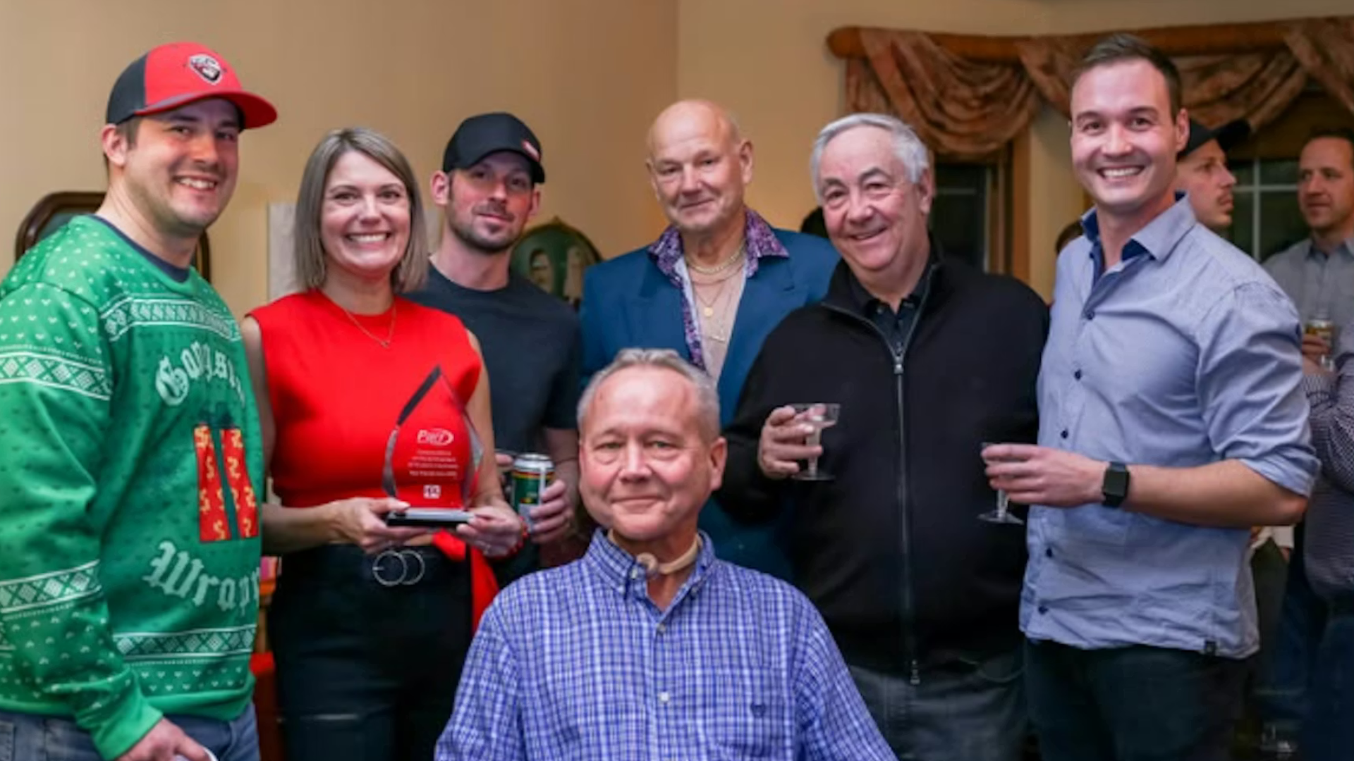 Group of seven people smiling at indoor event, one holding red award