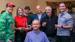 Group of seven people smiling at indoor event, one holding red award