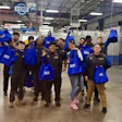 Team of automotive technicians celebrating in blue branded jackets at an industrial warehouse facility