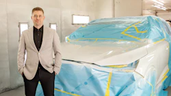 Professional man in beige blazer standing beside blue-wrapped vehicle in industrial workshop
