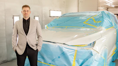 Professional man in beige blazer standing beside blue-wrapped vehicle in industrial workshop