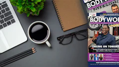 Workspace desk setup with laptop, coffee cup, notebook, and BODYWORX Professional magazine
