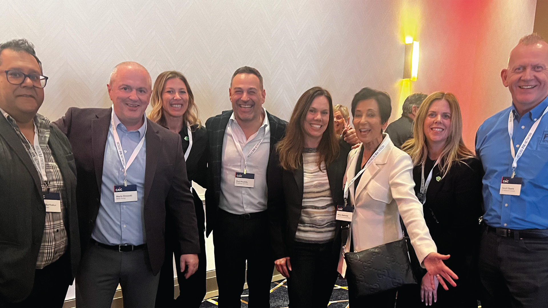 Group of nine professionals posing together at a corporate event wearing business attire and conference badges