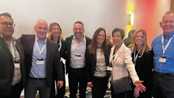 Group of nine professionals posing together at a corporate event wearing business attire and conference badges