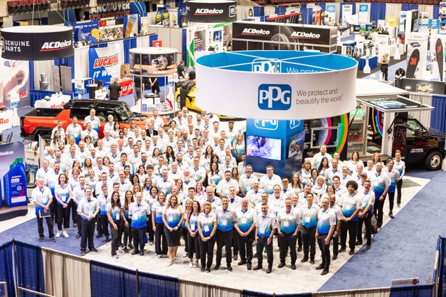 Large group of PPG employees in white and blue uniforms gathered around a branded cylindrical display at a corporate trade show or event