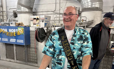 Man wearing turquoise Hawaiian shirt and sash smiling in industrial warehouse facility