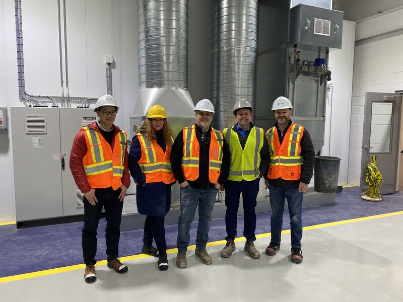 Five construction workers in hard hats and safety vests standing in an industrial facility with metal ductwork.