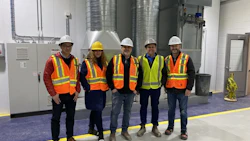 Five construction workers in hard hats and safety vests standing in an industrial facility with metal ductwork.