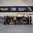 Large group of employees posing together in front of automotive service business with company logos on signage